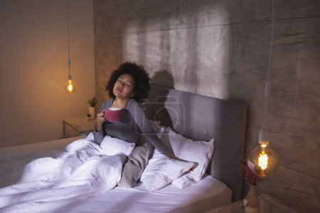 Photo for Beautiful young mixed race woman wearing pajamas lying in bed in the morning, drinking tea and enjoying leisure time at home - Royalty Free Image