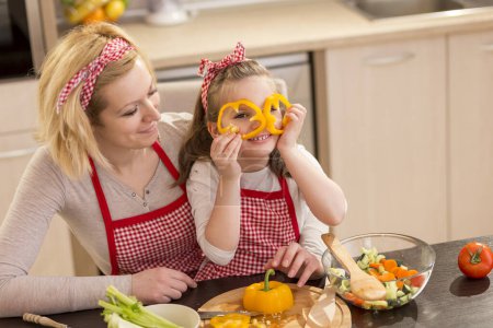 Téléchargez les photos : Belle mère et fille s'amusent à faire de la salade dans la cuisine, tenant des tranches de poivre comme verres et jetant un coup d'oeil. Focus sur la fille - en image libre de droit