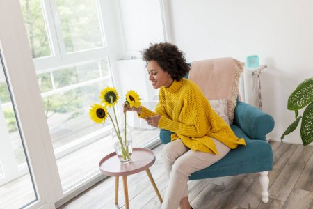 Foto de Hermosa joven mujer de raza mixta sentada en un sillón junto a la ventana, relajándose en casa, colocando ramo de girasol en un jarrón - Imagen libre de derechos