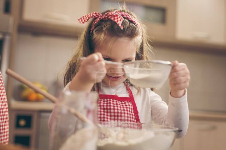 Foto de Niña ayudando a su madre en la cocina, sembrando harina a través del tamiz en un tazón de amasar - Imagen libre de derechos