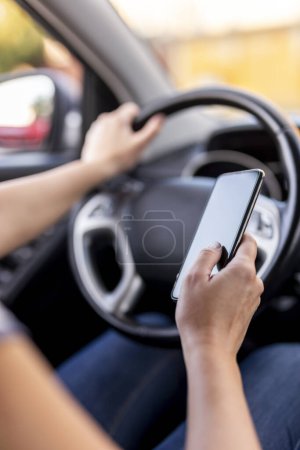 Foto de Detalle de las manos femeninas sosteniendo un volante y escribiendo un mensaje de texto en un teléfono inteligente mientras conduce un automóvil - Imagen libre de derechos