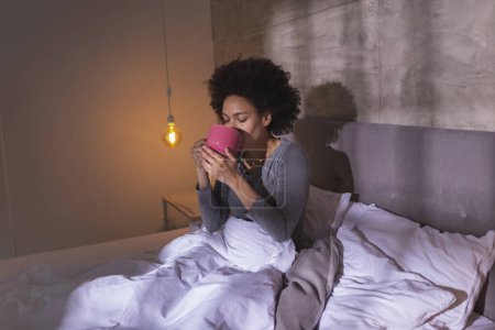 Foto de Hermosa joven mujer de raza mixta con pijama acostado en la cama por la mañana, bebiendo café y disfrutando del tiempo libre en casa - Imagen libre de derechos