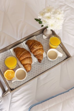 Téléchargez les photos : Vue de dessus d'un plateau de petit déjeuner avec vase de fleurs, jus d'orange, croissants et café placés sur le lit. Focus sélectif sur le croissant droit - en image libre de droit