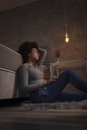 Photo for Beautiful young mixed race woman sitting on the floor by the bed with her eyes closed, tired after a long day, having a glass of wine - Royalty Free Image