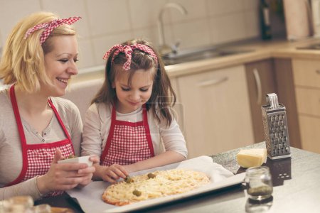 Photo for Mother and daughter making pizza in the kitchen; little girl putting olives on top of a pizza ready for baking. Focus on the daughter - Royalty Free Image