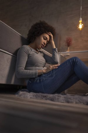 Photo for Beautiful young mixed race woman sitting on the floor by the bed with her eyes closed, tired after a long day, sad and stressed out - Royalty Free Image