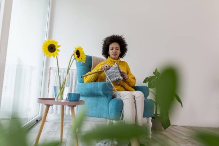 Photo for Beautiful young mixed race woman sitting in an armchair,  knitting a scarf and relaxing at home - Royalty Free Image