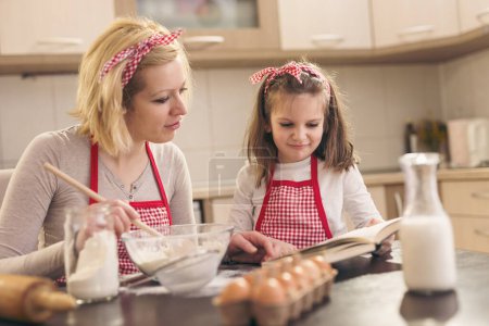 Photo for Mother and daughter making cookies in the kitchen, looking for a recipe in a cookbook - Royalty Free Image