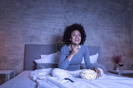 Foto de Hermosa mujer de raza mixta con pijamas sentados en la cama, comiendo palomitas de maíz y viendo una película de comedia en la televisión, relajándose en casa tarde en la noche - Imagen libre de derechos