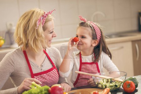 Foto de Madre e hija cortando verduras, haciendo ensalada y divirtiéndose; hija colocando tomate cherry como nariz - Imagen libre de derechos