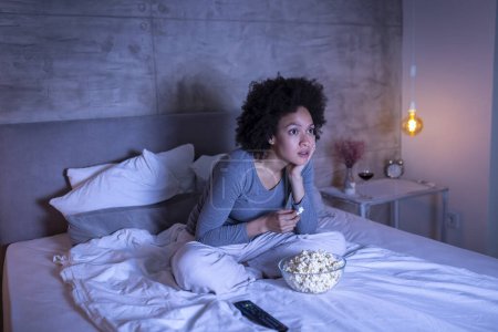 Téléchargez les photos : Belle femme mixte portant un pyjama assis sur le lit, mangeant du pop-corn et regardant un film d'horreur à la télévision, effrayée et tendue - en image libre de droit