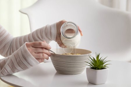 Téléchargez les photos : Détail des mains féminines versant du lait dans un bol de céréales ; femme prenant le petit déjeuner. Concentrez-vous sur les céréales et le bout de la bouteille - en image libre de droit