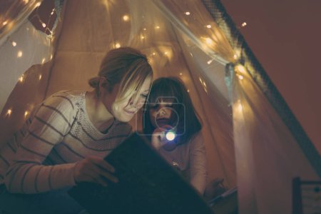 Photo for Mother and daughter sitting in a teepee, reading stories with the flashlight. Focus on the daughter - Royalty Free Image
