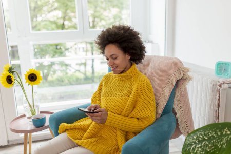 Foto de Hermosa joven mujer de raza mixta sentada en un sillón, relajándose en casa, charlando con amigos usando un teléfono inteligente - Imagen libre de derechos