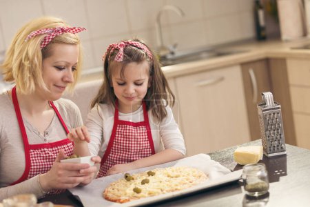 Téléchargez les photos : Mère et fille préparant des pizzas dans la cuisine ; petite fille mettant des olives sur une pizza prête à cuire. Focus sur la fille - en image libre de droit