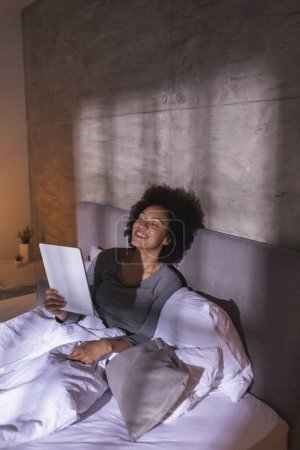 Téléchargez les photos : Belle jeune femme de race mixte portant un pyjama couché dans le lit le matin, ayant un appel vidéo à l'aide d'un ordinateur tablette et se détendre à la maison - en image libre de droit
