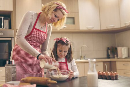 Téléchargez les photos : Pâte à pâtisserie mère et fille dans la cuisine ; mère ajoutant la farine d'un bocal pendant que la fille remuait avec une cuillère de cuisine - en image libre de droit
