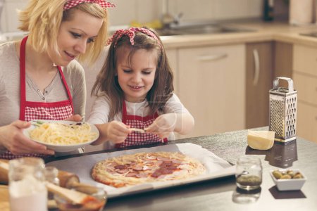 Foto de Madre e hija en la cocina haciendo pizza, poniendo el queso en la masa de pizza. Concéntrate en la hija - Imagen libre de derechos
