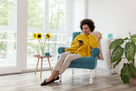 Téléchargez les photos : Belle femme métisse assise dans un fauteuil, relaxante à la maison, tapant un message texte sur un smartphone - en image libre de droit