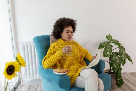 Photo for Beautiful mixed race woman relaxing at home, sitting in an armchair, reading newspapers and having breakfast - Royalty Free Image