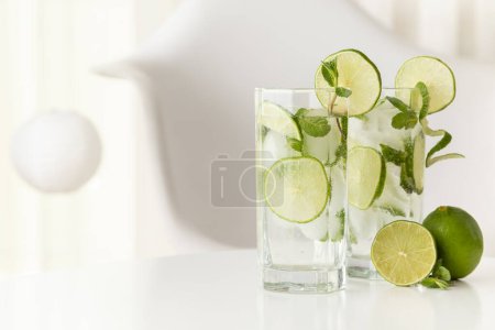 Foto de Dos cócteles mojito con mucho hielo, ron blanco, zumo de limón y tónico, decorados con rodajas de lima y hojas de menta en una moderna mesa blanca. - Imagen libre de derechos