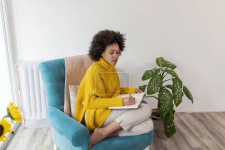 Photo for Portrait of a beautiful mixed race woman sitting in an armchair, taking notes in a planner - Royalty Free Image
