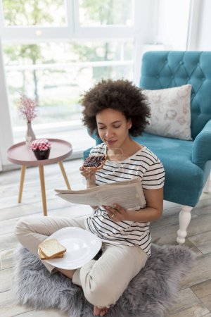 Foto de Hermosa mujer de raza mixta relajándose en casa, leyendo periódicos y desayunando - Imagen libre de derechos