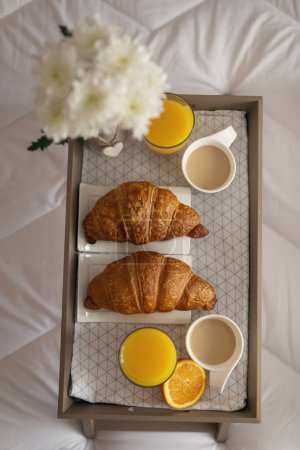 Téléchargez les photos : Vue de dessus d'un plateau de petit déjeuner avec vase de fleurs, jus d'orange, croissants et café placés sur le lit. Focus sur le croissant supérieur - en image libre de droit