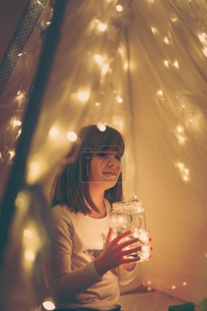 Foto de Chica jugando en un tipi, sosteniendo un frasco lleno de luces de Navidad - Imagen libre de derechos