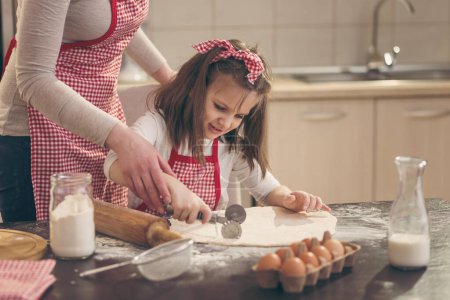 Foto de Hermosa niña con delantal en la cocina cortando masa para rollos con cortador de pizza con la ayuda de su madre - Imagen libre de derechos