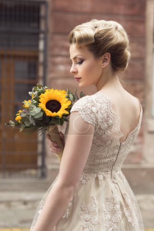 Photo for Beautiful young bride posing in a wedding dress in a retro cobble street, holding a sunflower bouquet - Royalty Free Image