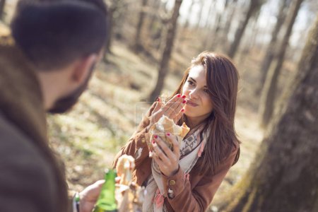 Téléchargez les photos : Jeune couple assis sur un banc en bois au bord de la route forestière, faisant une pause en mangeant des sandwichs et en buvant de la bière - en image libre de droit