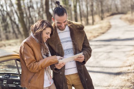 Téléchargez les photos : Couple faisant une pause sur la route, se tenant près de la route et analysant leur itinéraire sur un système de navigation tablette - en image libre de droit