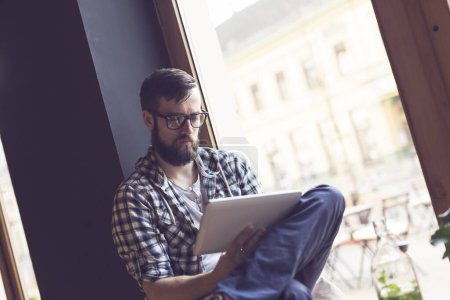 Téléchargez les photos : Homme barbu hipster assis à côté d'une fenêtre dans un café de design d'intérieur moderne, travaillant sur une tablette - en image libre de droit