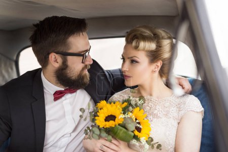 Téléchargez les photos : Jeune couple de jeunes mariés assis dans une voiture vintage rétro, embrasser et partir en lune de miel - en image libre de droit