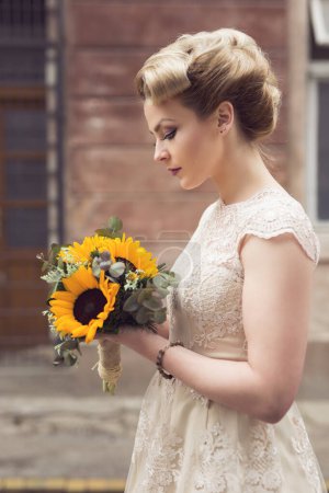 Téléchargez les photos : Belle jeune mariée posant dans une robe de mariée dans une rue pavée rétro, tenant un bouquet de tournesol - en image libre de droit
