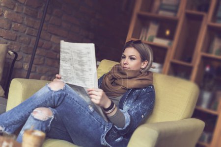 Téléchargez les photos : Belle jeune femme assise dans un fauteuil dans un café, lisant les journaux - en image libre de droit
