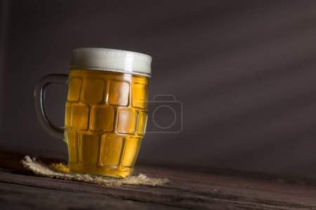 Téléchargez les photos : Tasse de bière froide et pâle placée sur une couverture de toile de jute sur une table en bois rustique - en image libre de droit