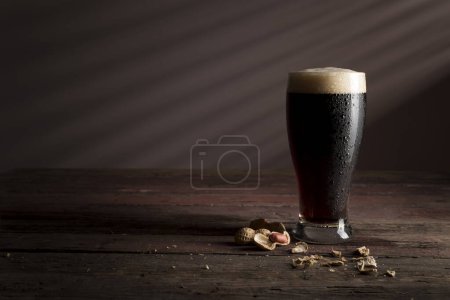 Foto de Vaso de cerveza fría oscura con espuma colocado sobre una mesa rústica de madera con unos cacahuetes al lado - Imagen libre de derechos