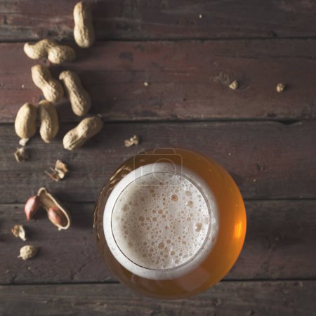 Foto de Vista superior de un vaso de cerveza pálida sin filtrar con espuma y algunos cacahuetes en una mesa de pub de madera rústica. Concéntrate en la espuma - Imagen libre de derechos