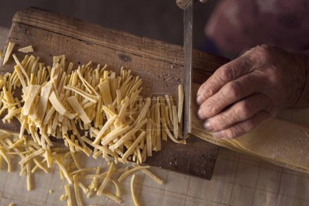 Photo for Top view of an elderly woman's hand cutting noodles with an old knife while making homemade pasta. Selective focus - Royalty Free Image