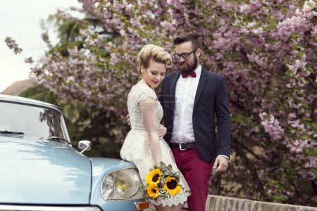 Téléchargez les photos : Couple nouvellement marié debout dans la rue à côté d'une vieille voiture rétro, embrasser et embarquer sur une lune de miel - en image libre de droit
