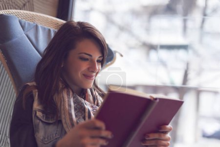 Téléchargez les photos : Belle jeune femme assise dans un café à côté d'une fenêtre, souriant et lisant un livre - en image libre de droit