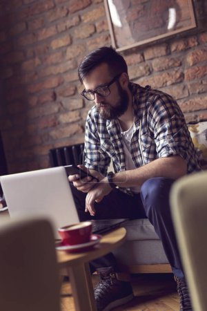 Téléchargez les photos : Beau mec hipster assis dans un fauteuil dans un café, prenant un café du matin et utilisant un ordinateur portable et un téléphone intelligent pour son travail indépendant - en image libre de droit