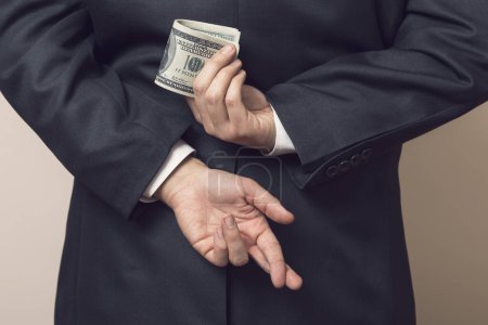 Foto de Empresario en un traje sosteniendo sus manos detrás de su espalda con una pila de billetes de dólar en uno y los dedos cruzados en otro - Imagen libre de derechos