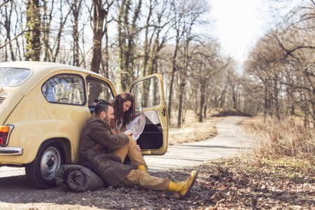 Téléchargez les photos : Jeune couple amoureux faisant une pause sur leur route, assis au bord de la route et regardant une carte, essayant de décider de leur itinéraire. Concentre-toi sur la fille - en image libre de droit