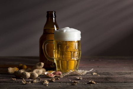 Téléchargez les photos : Bouteille de bière et une tasse de bière froide et pâle placée sur une couverture de toile de jute avec quelques cacahuètes sur une table en bois rustique - en image libre de droit