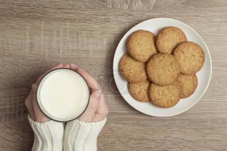 Foto de Vista superior de las manos femeninas sosteniendo un vaso de leche, con el plato de galletas de chispas de chocolate colocado junto a ella en la mesa - Imagen libre de derechos