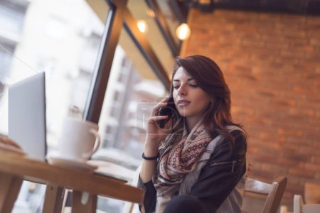 Téléchargez les photos : Jolie jeune brune assise dans un café et ayant une conversation téléphonique - en image libre de droit