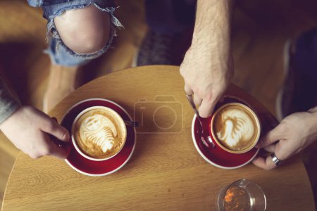 Foto de Vista superior de dos personas que sostienen una taza de café de arte latte bien decorado, disfrutando de su café de la mañana. Concéntrate en el café de la izquierda - Imagen libre de derechos